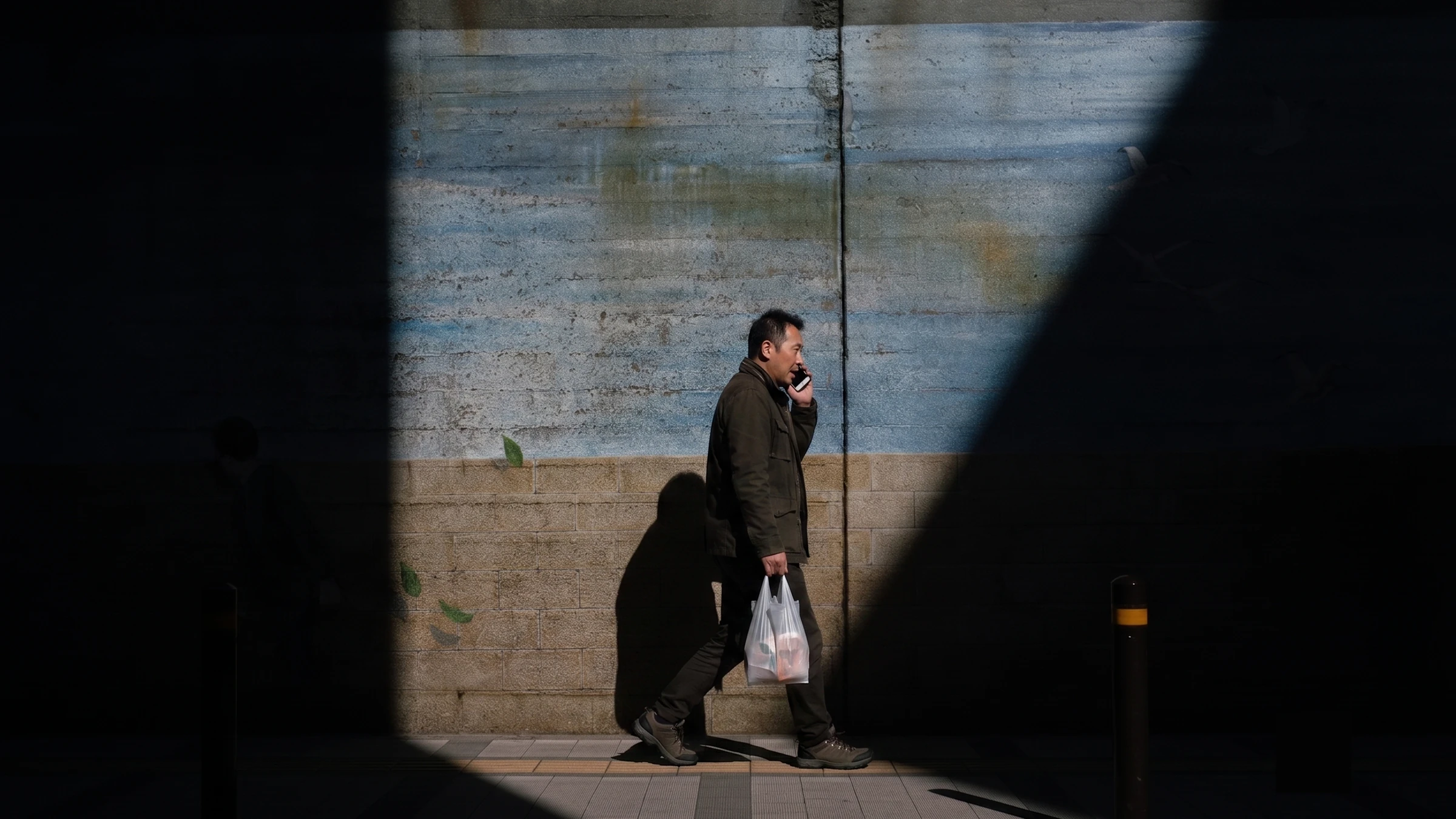 A man mid-stride on a sunlit street, talking on the phone.